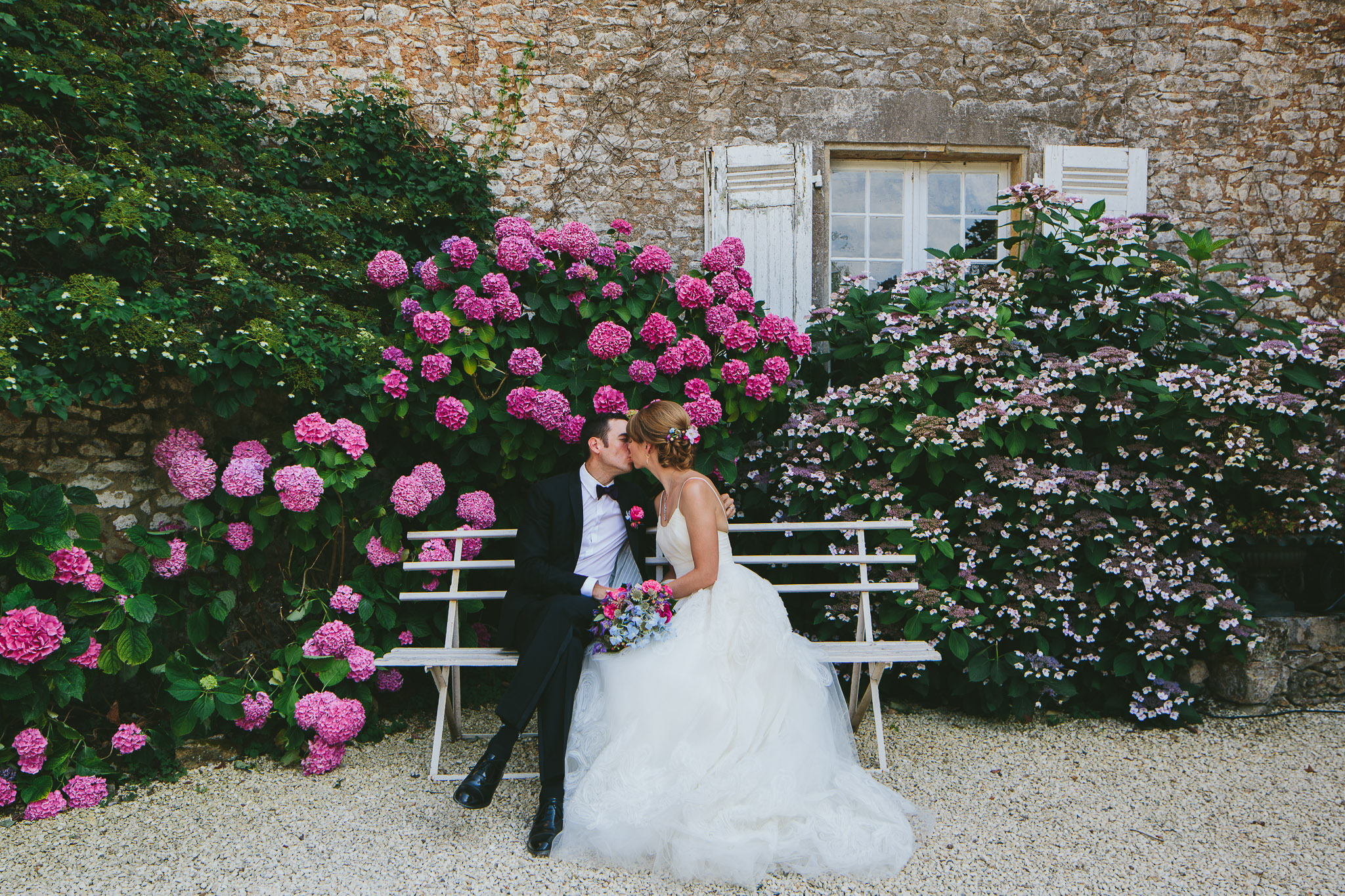 French professional wedding photographer newly weds kissing on a bench with pink hydrangeas in the background